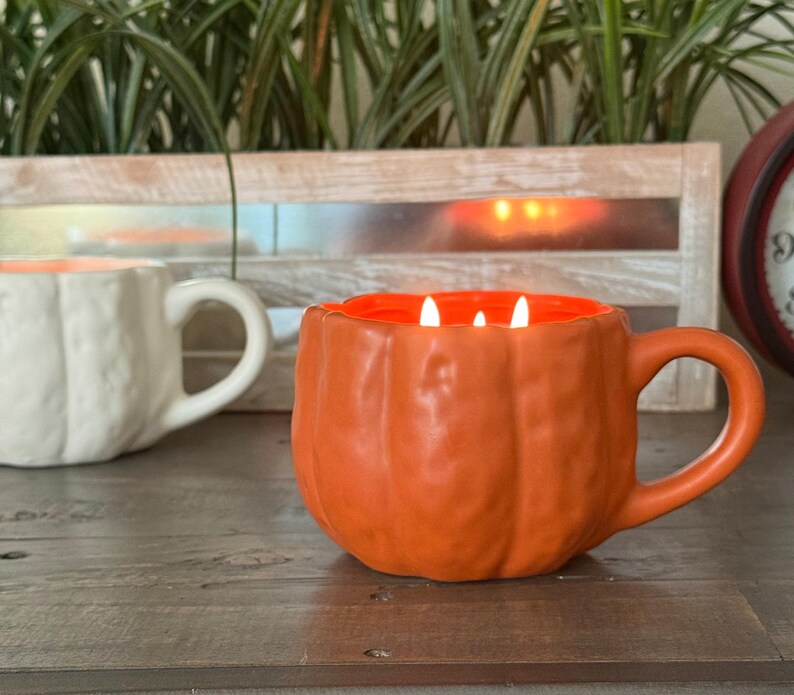 May include: An orange pumpkin-shaped mug candle with three lit wicks. The interior of the mug is orange, and the exterior has a textured pumpkin design. A white pumpkin-shaped mug is in the background. The mugs are on a wooden surface.