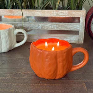 May include: An orange pumpkin-shaped candle with three lit wicks, resembling a mug. The candle sits on a wooden surface, with a white mug-shaped candle in the background. The candle has a handle and is filled with orange wax.