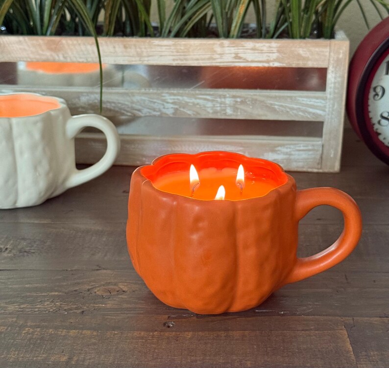 May include: An orange pumpkin-shaped candle with three lit wicks, resembling a mug. The candle sits on a wooden surface, with a white mug-shaped candle in the background. The candle has a handle and is filled with orange wax.