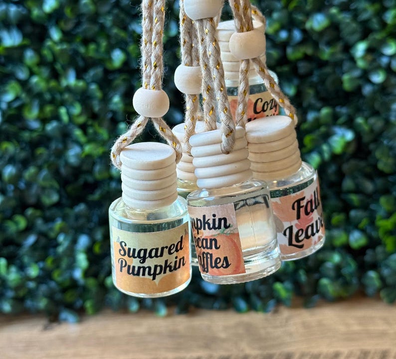 Puede incluir: Una colecci&oacute;n de peque&ntilde;as botellas de vidrio transparente con tapas de madera y etiquetas decorativas. Las etiquetas presentan textos como "Sugared Pumpkin" y "Fall Leaves". Cada botella est&aacute; suspendida por una cuerda, lo que sugiere que son ambientadores.