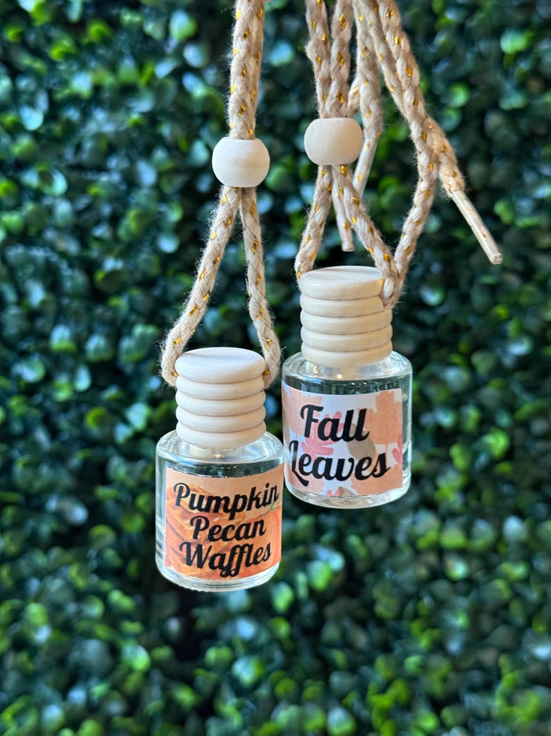 Puede incluir: Dos peque&ntilde;os frascos de vidrio con tapas de madera y etiquetas. Una etiqueta dice "Pumpkin Pecan Waffles" y la otra "Fall Leaves". Los frascos est&aacute;n suspendidos por una cuerda dorada con cuentas de madera, sobre un fondo verde.