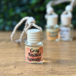 Puede incluir: Una peque&ntilde;a botella de vidrio con tapa de madera y una etiqueta que dice "Cozy Knitted Sweater". La botella est&aacute; suspendida por una cuerda beige con detalles dorados. El fondo est&aacute; borroso, con toques de follaje verde y otras botellas.
