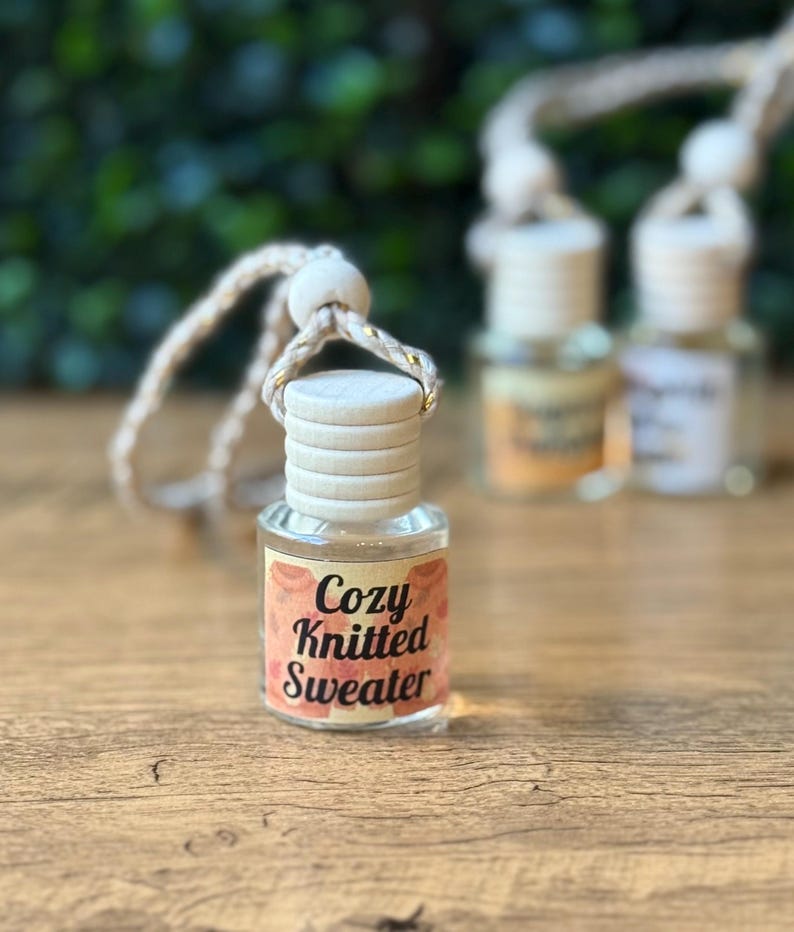 Puede incluir: Una peque&ntilde;a botella de vidrio con tapa de madera y una etiqueta que dice "Cozy Knitted Sweater". La botella est&aacute; suspendida por una cuerda beige con detalles dorados. El fondo est&aacute; borroso, con toques de follaje verde y otras botellas.