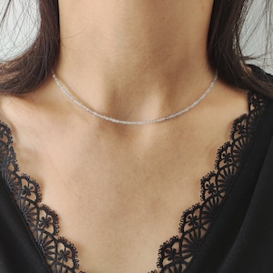 Puede incluir: Un delicado collar de plata con pequeñas cuentas de color gris claro. El collar lo lleva una persona con el pelo oscuro y una blusa de encaje negra.