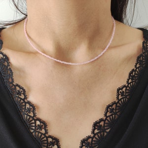 Puede incluir: Un delicado collar de cuentas rosas. El collar se lleva sobre una blusa negra con un ribete de encaje decorativo. El collar parece estar hecho de peque&ntilde;as cuentas redondas, creando un accesorio sutil y elegante.