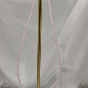 Puede incluir: Un delicado collar con peque&ntilde;as cuentas de color rosa claro. El collar se exhibe sobre un soporte dorado sobre un fondo blanco. Las cuentas son de tama&ntilde;o uniforme y est&aacute;n muy juntas, creando un aspecto sutil y elegante.