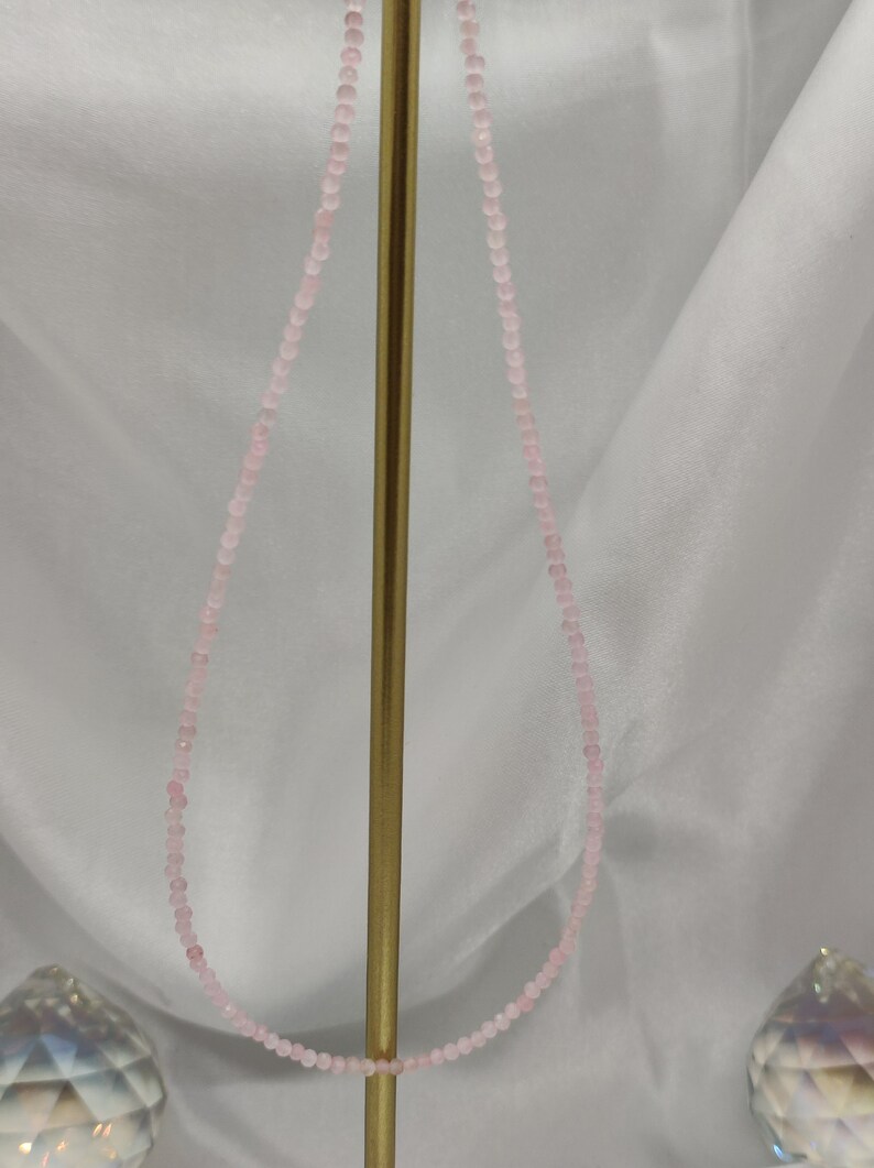 Puede incluir: Un delicado collar con peque&ntilde;as cuentas de color rosa claro. El collar se exhibe sobre un soporte dorado sobre un fondo blanco. Las cuentas son de tama&ntilde;o uniforme y est&aacute;n muy juntas, creando un aspecto sutil y elegante.