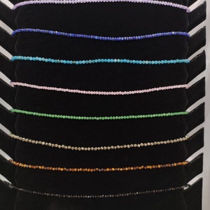 Puede incluir: Exhibici&oacute;n de delicados collares de cuentas en varios colores, incluyendo lavanda, azul, turquesa, rosa, verde, dorado, marr&oacute;n y negro. Cada collar est&aacute; compuesto por peque&ntilde;as cuentas espaciadas uniformemente, creando un brillo sutil. Los collares se presentan sobre un expositor de terciopelo negro.