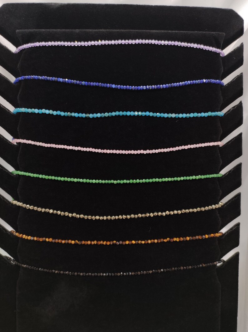 Puede incluir: Exhibici&oacute;n de delicados collares de cuentas en varios colores, incluyendo lavanda, azul, turquesa, rosa, verde, dorado, marr&oacute;n y negro. Cada collar est&aacute; compuesto por peque&ntilde;as cuentas espaciadas uniformemente, creando un brillo sutil. Los collares se presentan sobre un expositor de terciopelo negro.