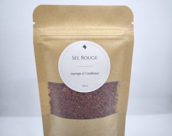 May include: A brown paper bag with a clear window displaying red salt crystals. The bag has a white circular label with the text "SEL ROUGE" and "Ancrage & Confiance". The bag is filled with 80g of salt.