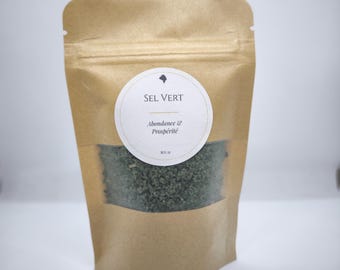 May include: A brown paper bag with a clear window displaying green sea salt. A white circular label on the bag reads "SEL VERT" with the words "Abondance & Prospérité" below. The bag is sealed at the top.