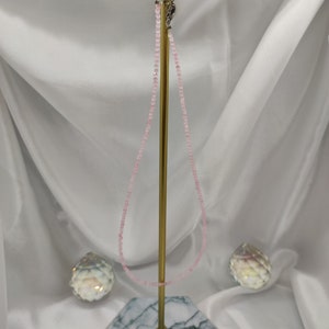 Puede incluir: Un delicado collar de cuentas rosas expuesto sobre un soporte dorado con base de m&aacute;rmol. El collar est&aacute; compuesto por peque&ntilde;as cuentas redondas. Dos objetos decorativos de cristal a cada lado del soporte.