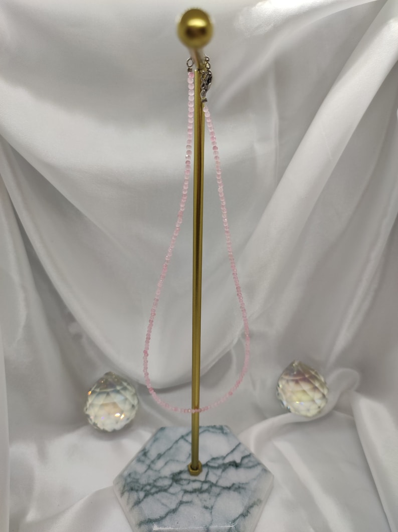 Puede incluir: Un delicado collar de cuentas rosas expuesto sobre un soporte dorado con base de m&aacute;rmol. El collar est&aacute; compuesto por peque&ntilde;as cuentas redondas. Dos objetos decorativos de cristal a cada lado del soporte.