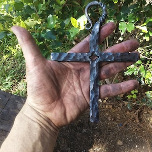 Pode incluir: Uma cruz de metal forjada à mão com acabamento martelado e um laço de metal torcido no topo. A cruz é cinza escura e tem um pequeno furo no centro.