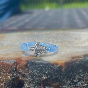 May include: A light blue beaded ring with a silver turtle charm. The ring is on a brown and white stone surface.