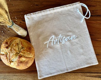 May include: A rustic still life featuring a freshly baked loaf of bread next to a beige drawstring bag with the word "Artisan" in white script. A small glass bottle with wheat stalks and grains is also present.
