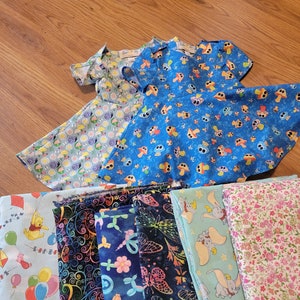 May include: A collection of fabric scraps in various patterns and colors, including blue, pink, green, and black. The fabrics feature whimsical designs such as Winnie the Pooh, butterflies, and mushrooms. There are also two blue dresses with different patterns.