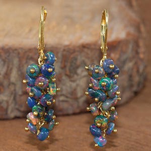 May include: A pair of gold-colored leverback earrings. Each earring features a cluster of iridescent blue, green, and pink opal beads. The opal beads are connected with small gold-colored accents.