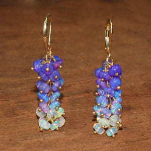 May include: A pair of gold dangle earrings with a gradient of purple and white opal beads. The earrings are made with a delicate wire design and have a hook closure.