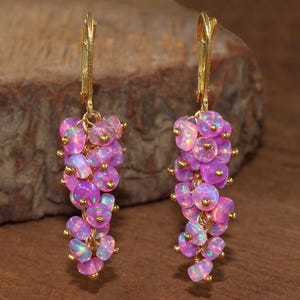 Può includere: Un paio di orecchini pendenti con grappoli di opali rosa iridescenti. Gli opali sono attaccati a ganci per orecchie color oro. Gli orecchini hanno un design a cascata, con ogni grappolo di opali appeso a una piccola perla d'oro.