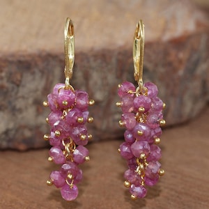 Op de afbeelding: Een paar oorhangers met trossen roze edelstenen, bevestigd aan gouden oorhaken. De edelstenen zijn helderroze, tegen een vage houten achtergrond.