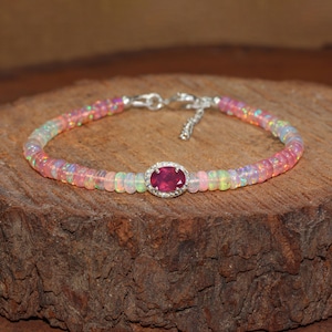 May include: A delicate bracelet featuring a central oval ruby surrounded by small diamonds. The bracelet's band is composed of small, iridescent opal beads in shades of pink, white, and yellow. The clasp is silver.