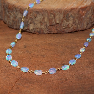May include: A delicate necklace featuring oval opals in shades of blue and white, strung on a gold-coloured chain. The opals have a subtle iridescence, displayed on a wooden surface.