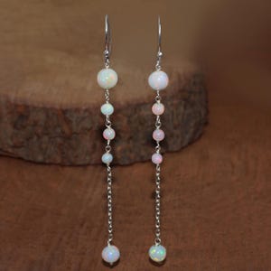 Könnte beinhalten: Hängeohrringe mit einer Reihe von opalisierenden, runden Perlen in verschiedenen Größen. Die Perlen sind an einer filigranen Silberkette aufgereiht, mit einer größeren Perle oben und unten. Die Ohrringe haben einen Hakenverschluss. Der Hintergrund ist ein verschwommenes, warmes Braun.