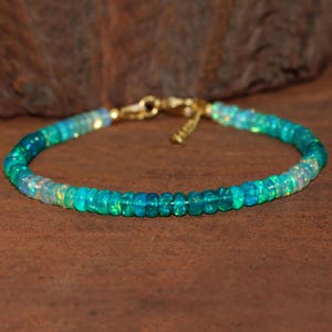 Könnte beinhalten: Ein Armband mit schillernden Opalsteinen in Türkis-, Grün- und Blautönen. Das Armband ist an einer zarten Goldkette mit passendem Verschluss befestigt. Die Edelsteine sind rund und gleichmäßig groß.