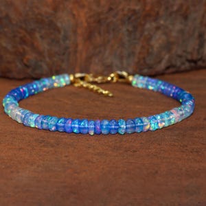 May include: A beaded bracelet featuring iridescent blue and white opal stones. The bracelet has a gold-colored chain and clasp. The opal beads are round and vary in shades of blue and white, creating a gradient effect.