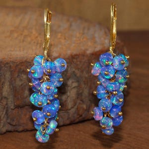 May include: A pair of gold-tone leverback earrings featuring clusters of blue opal beads. The opals have iridescent flashes of color, and are accented with small gold-colored beads. The earrings hang from gold-colored leverback closures.