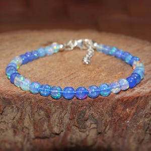 May include: A beaded bracelet with iridescent blue and white opal beads. The bracelet has a silver-coloured clasp and is displayed on a wooden surface. The beads are approximately 0.6 cm in diameter.