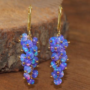 May include: A pair of gold-colored leverback earrings. Each earring features a cluster of iridescent blue opal beads with gold-colored accents. The earrings are displayed against a wooden background.