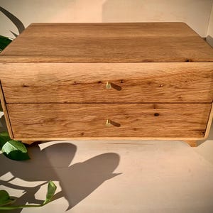May include: A rectangular wooden chest with two horizontal drawers, each featuring a small, circular brass knob. The chest is crafted from light brown oak wood with a visible natural grain pattern and sits on four short, integrated wooden feet.