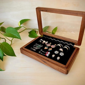 May include: A rectangular wooden jewelry box with a hinged glass lid and a black velvet interior lining. The box contains various earrings and rings arranged on the black surface, showcasing a natural wood grain finish and a clear glass top.