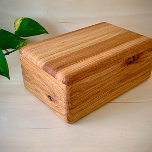 May include: A rectangular wooden box with a natural light brown grain pattern and rounded edges. The box features visible wood knots on the front and top surfaces, showcasing a smooth, polished finish with a distinct, organic texture throughout.