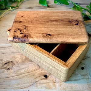 May include: A rectangular wooden box with a natural grain finish and a removable lid. The lid features prominent dark knots and wood grain patterns. The interior of the box is divided into two open compartments, showcasing the light brown wood construction.