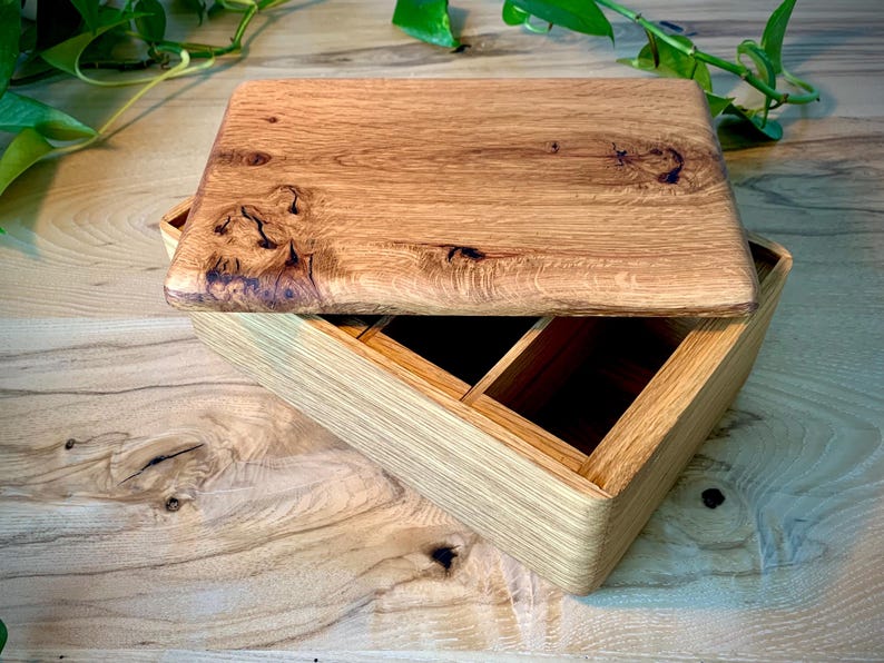 May include: A rectangular wooden box with a natural grain finish and a removable lid. The lid features prominent dark knots and wood grain patterns. The interior of the box is divided into two open compartments, showcasing the light brown wood construction.