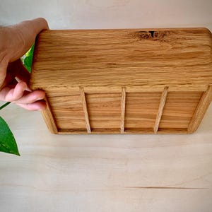 May include: A rectangular wooden box crafted from light brown oak with a visible natural wood grain pattern. The box features rounded corners and three vertical wooden slats attached to the front panel, showcasing a smooth, polished finish.