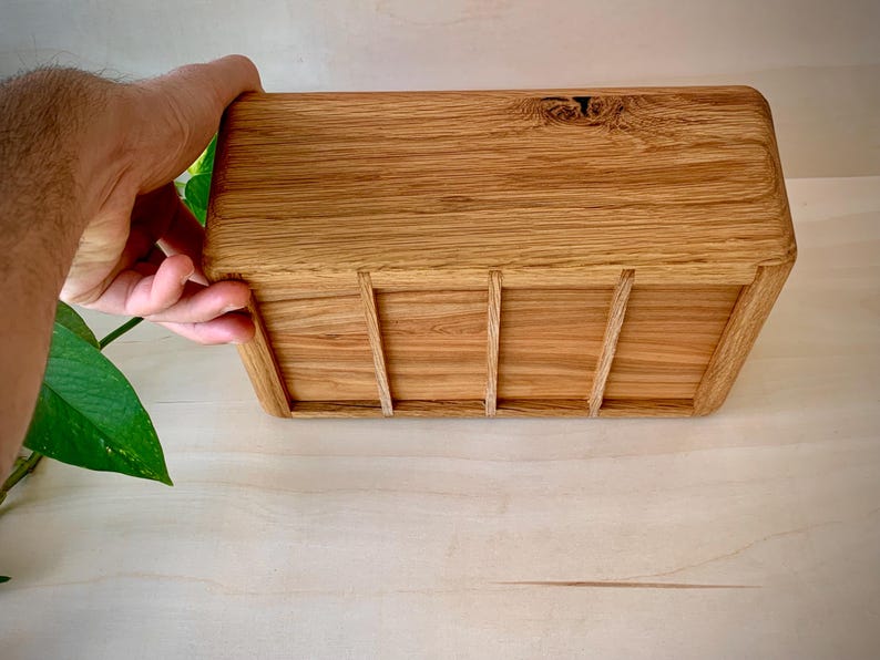 May include: A rectangular wooden box crafted from light brown oak with a visible natural wood grain pattern. The box features rounded corners and three vertical wooden slats attached to the front panel, showcasing a smooth, polished finish.