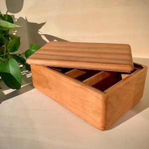 May include: A wooden box with a lid slightly ajar, revealing interior compartments. The box is made of light-colored wood with a reddish-brown lid. The box is sitting on a light-colored surface, with a green plant in the background.