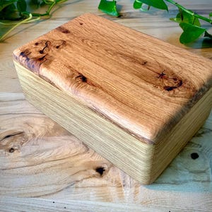 May include: A rectangular wooden box featuring a natural light brown finish with visible wood grain patterns. The lid displays prominent dark knots and swirling wood textures, while the sides show a layered, linear grain structure in a smooth finish.
