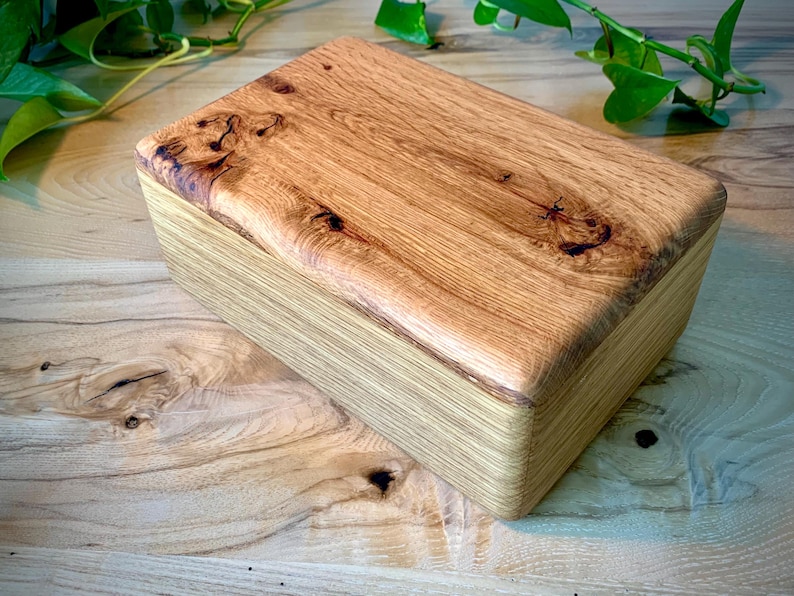 May include: A rectangular wooden box featuring a natural light brown finish with visible wood grain patterns. The lid displays prominent dark knots and swirling wood textures, while the sides show a layered, linear grain structure in a smooth finish.