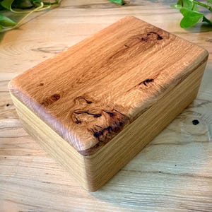 May include: A rectangular wooden box with a natural light brown finish. The lid features a prominent wood grain pattern with dark, organic knots and swirls, while the sides of the box display a smooth, layered, and straight-grained texture.