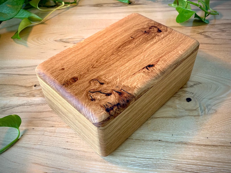 May include: A rectangular wooden box with a natural light brown finish. The lid features a prominent wood grain pattern with dark, organic knots and swirls, while the sides of the box display a smooth, layered, and straight-grained texture.
