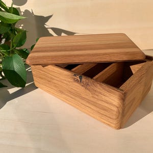 May include: A rectangular wooden box with a natural grain finish and a matching lid. The interior of the box is divided into four separate compartments by thin wooden dividers. The exterior features a visible knot in the wood grain on the front side.