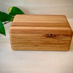 May include: A rectangular wooden box with a natural light brown finish and visible wood grain patterns. The box features rounded edges and a prominent knot detail on the front panel, showcasing the organic texture of the wood material.