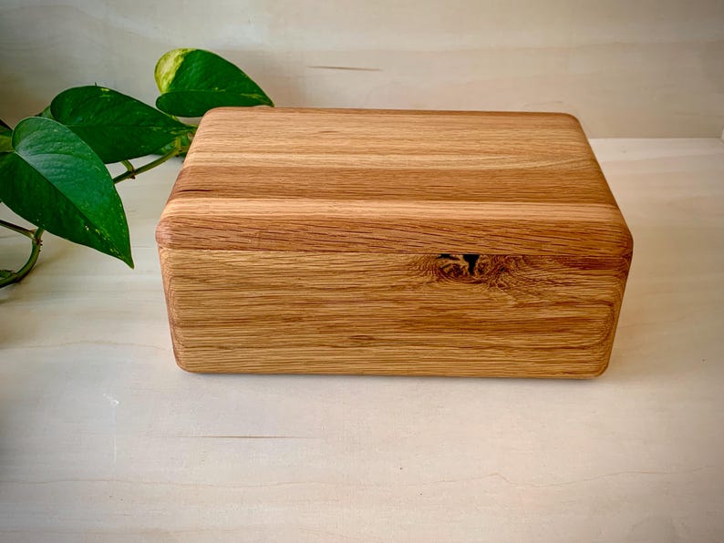 May include: A rectangular wooden box with a natural light brown finish and visible wood grain patterns. The box features rounded edges and a prominent knot detail on the front panel, showcasing the organic texture of the wood material.