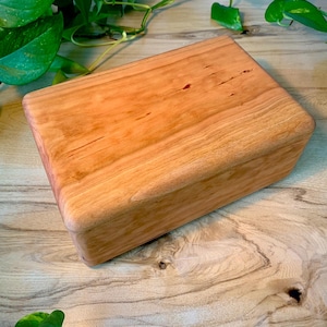 May include: A rectangular wooden box with a smooth, light brown finish and visible natural grain patterns. The box features rounded edges and a seamless lid design, showcasing the organic texture and warm tones of the polished wood surface.