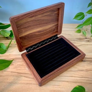 May include: A rectangular wooden jewelry box with a hinged lid, featuring a rich brown wood grain finish. The interior base is lined with a black velvet insert containing multiple parallel slots designed for organizing rings or small accessories.
